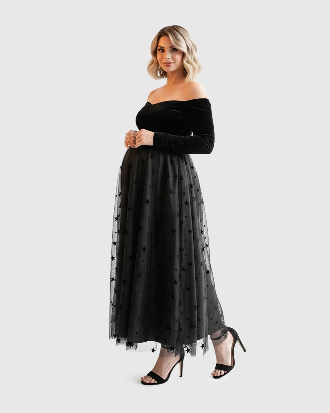 Maternity midi dress: black velvet off-shoulder bodice, star-flocked mesh overlay skirt, worn by a smiling pregnant woman.