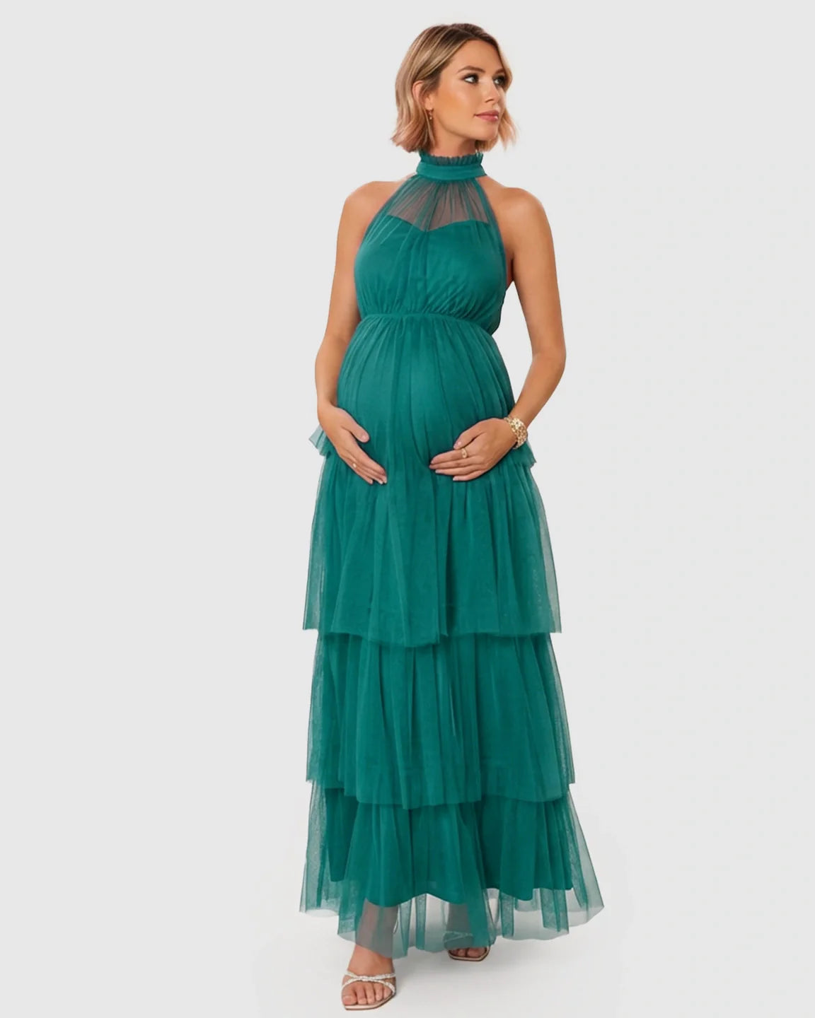 Maternity model in a teal halter-neck tiered mesh gown, hands cradling her bump.