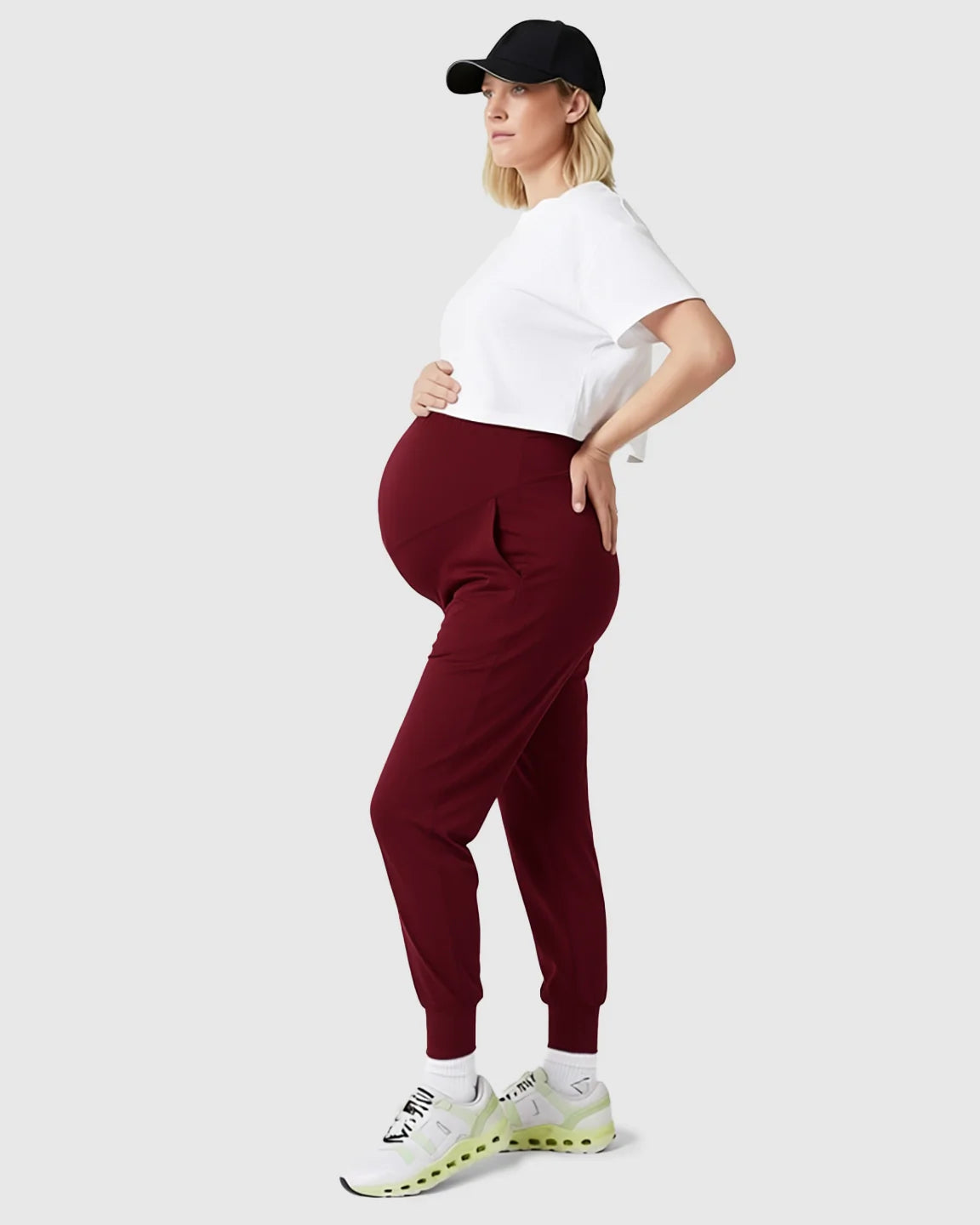 Pregnant woman in a white tee, black cap, and supportive slim-fitting burgundy maternity joggers.