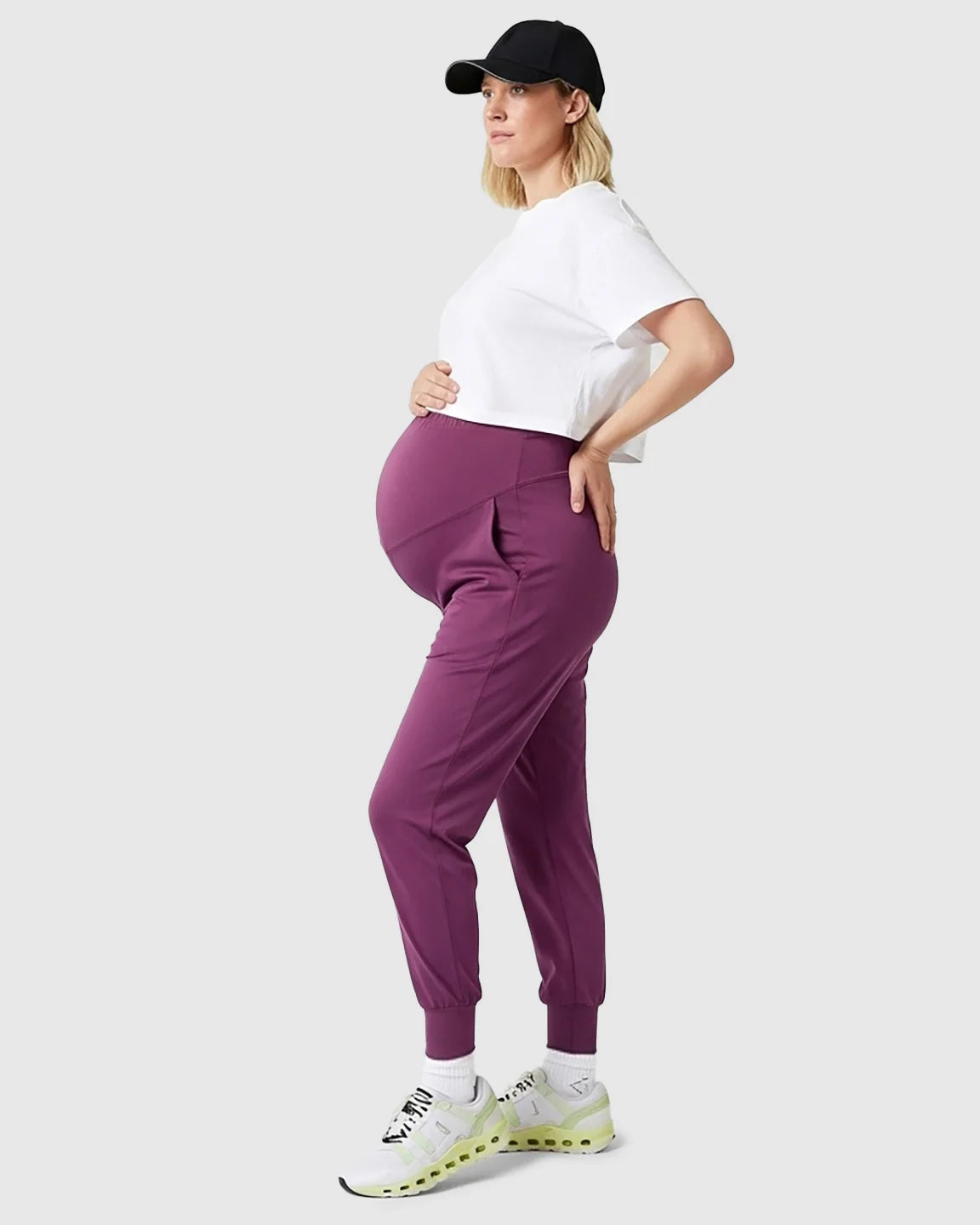 Pregnant woman in supportive plum maternity joggers, white crop top, and black cap.
