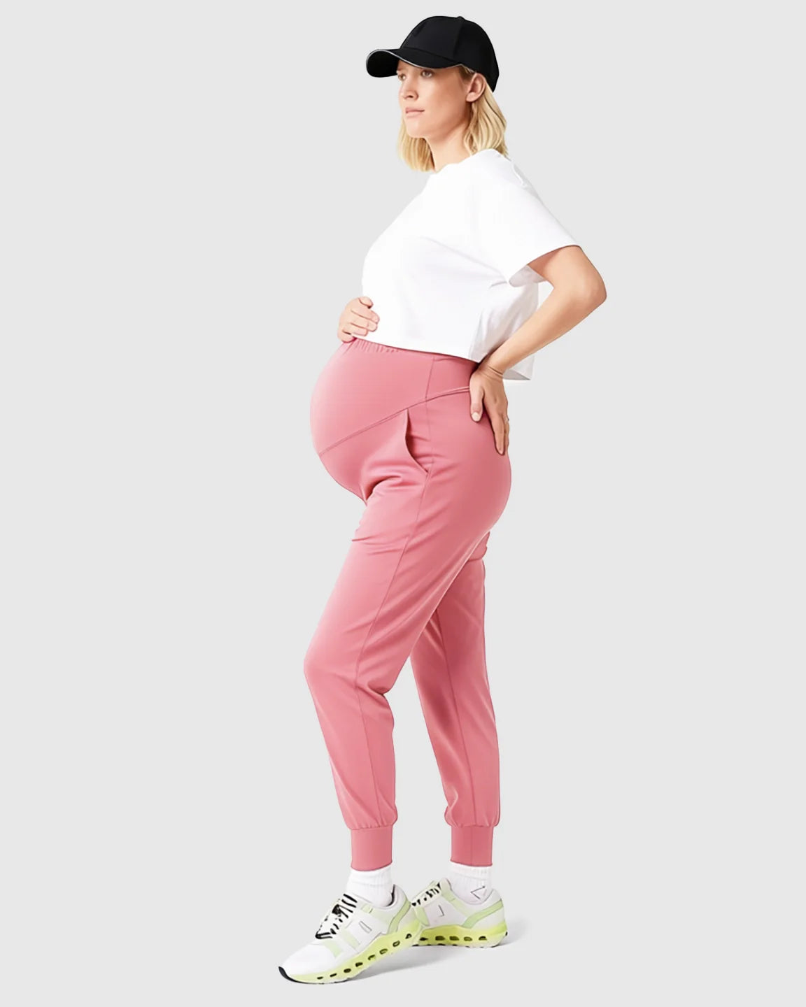 Pregnant woman in pink supportive slim-fitting maternity joggers, white crop top, and black cap.