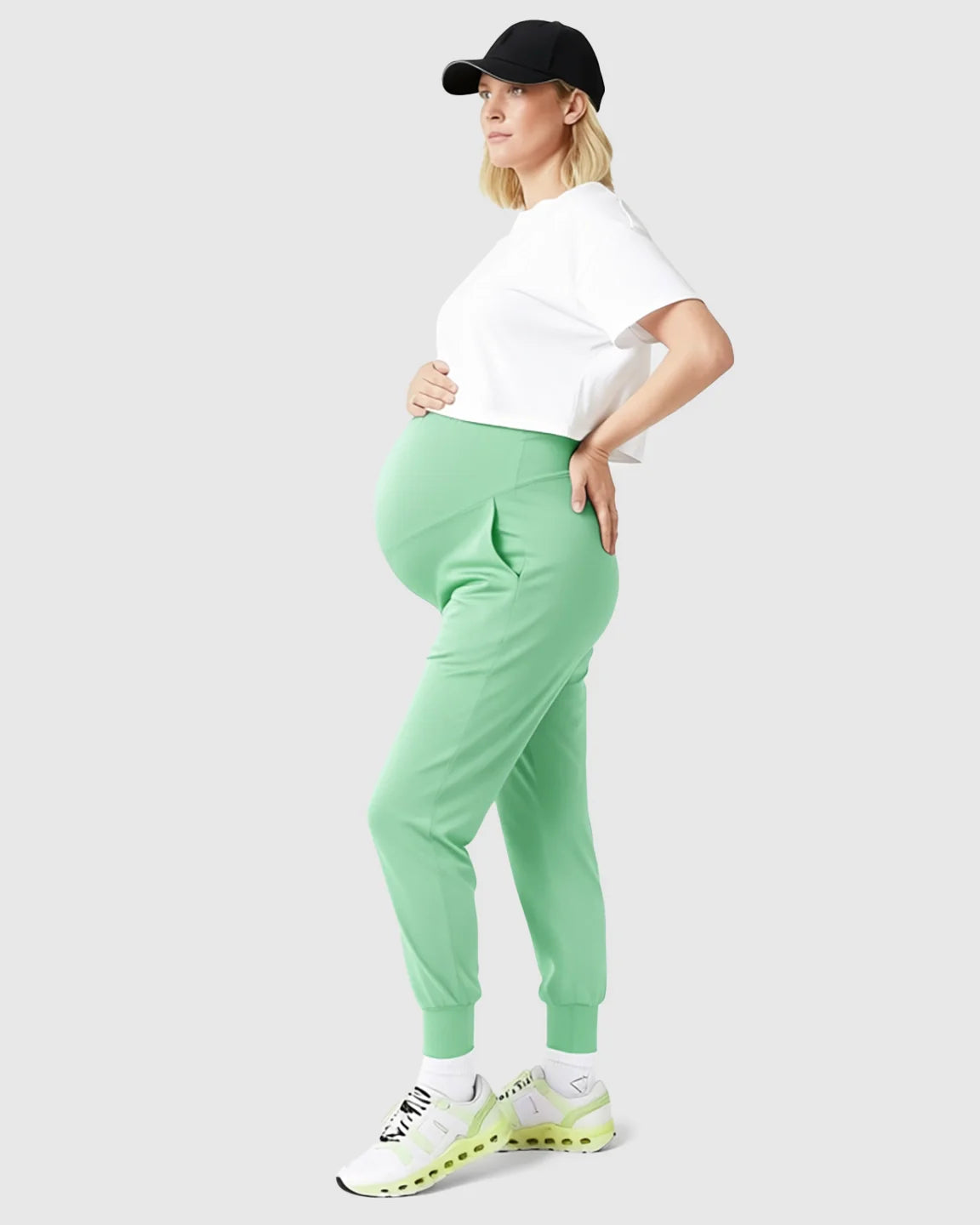 Pregnant woman in light green supportive slim-fitting maternity joggers, white crop top, and black cap.