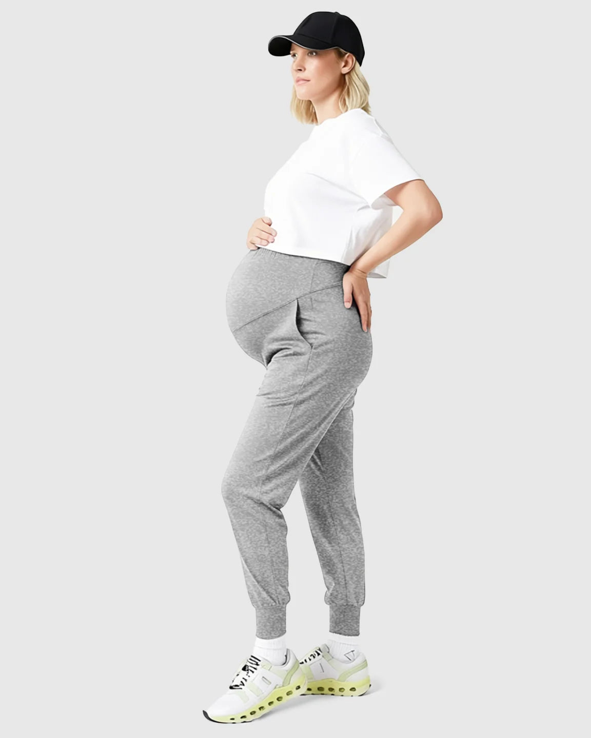 Pregnant woman in gray supportive slim-fitting maternity joggers, white crop top, black cap, and sneakers.