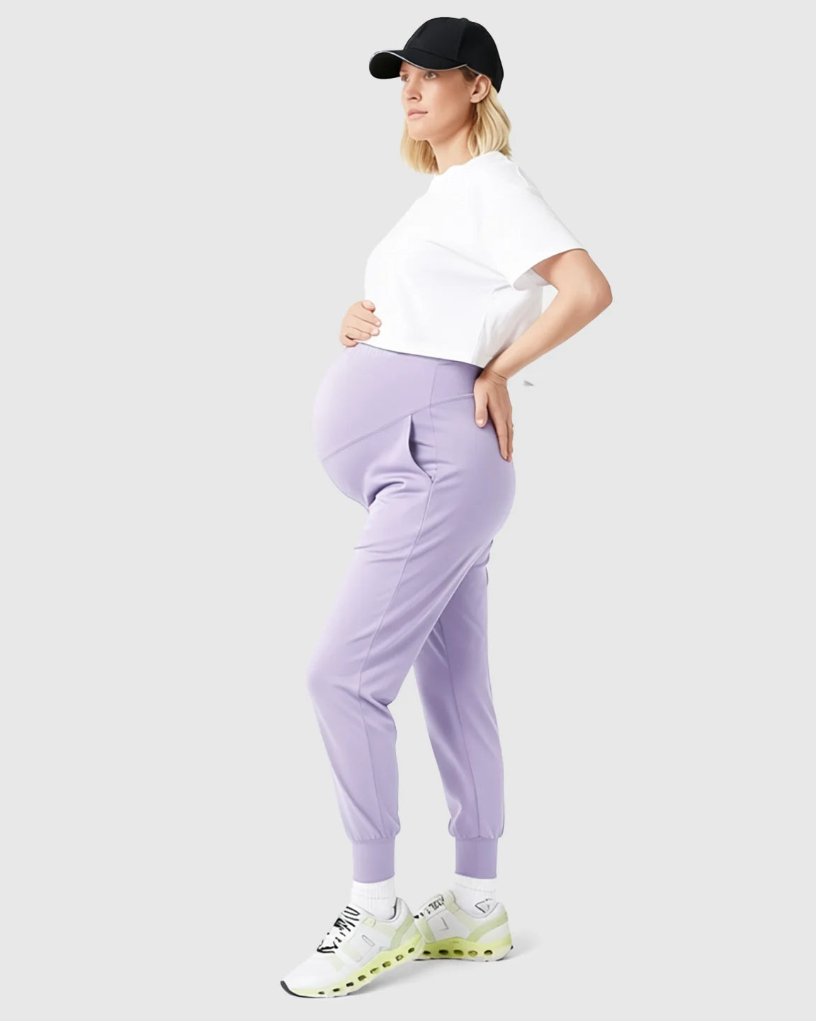Pregnant woman in lavender supportive slim-fitting maternity joggers, white tee, black cap, and sneakers.
