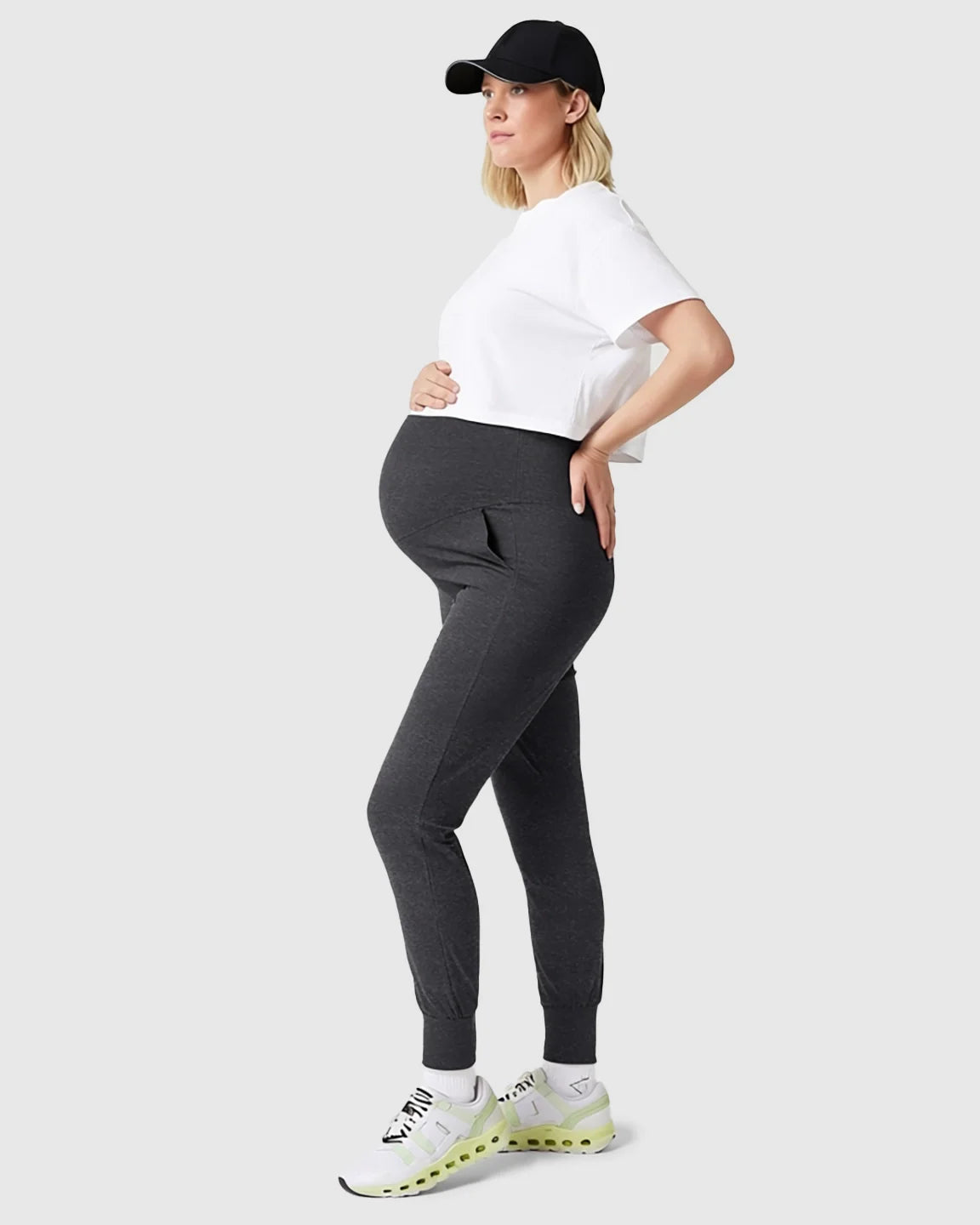 Pregnant woman in dark gray supportive slim-fitting maternity joggers with a high-waist and white crop top.