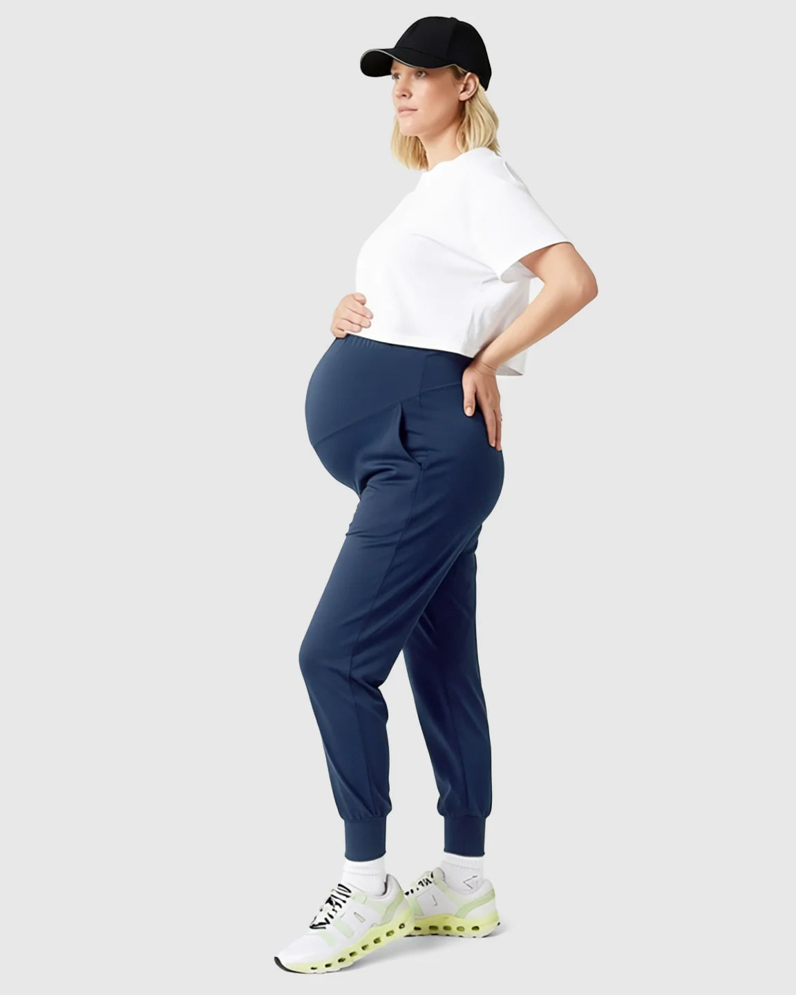 Pregnant woman in navy supportive slim-fitting maternity joggers, white crop top, and black cap.