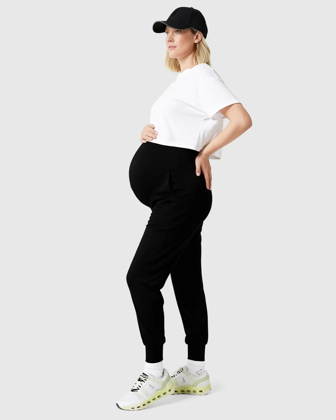 Pregnant woman in black supportive slim-fitting maternity joggers, white crop top, and black baseball cap.