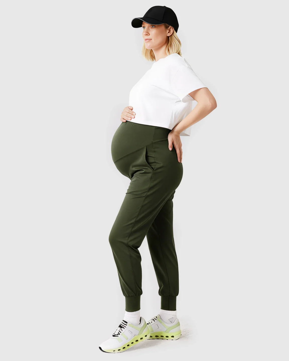 Pregnant woman in olive green supportive slim-fitting maternity joggers, white crop top, and black cap.