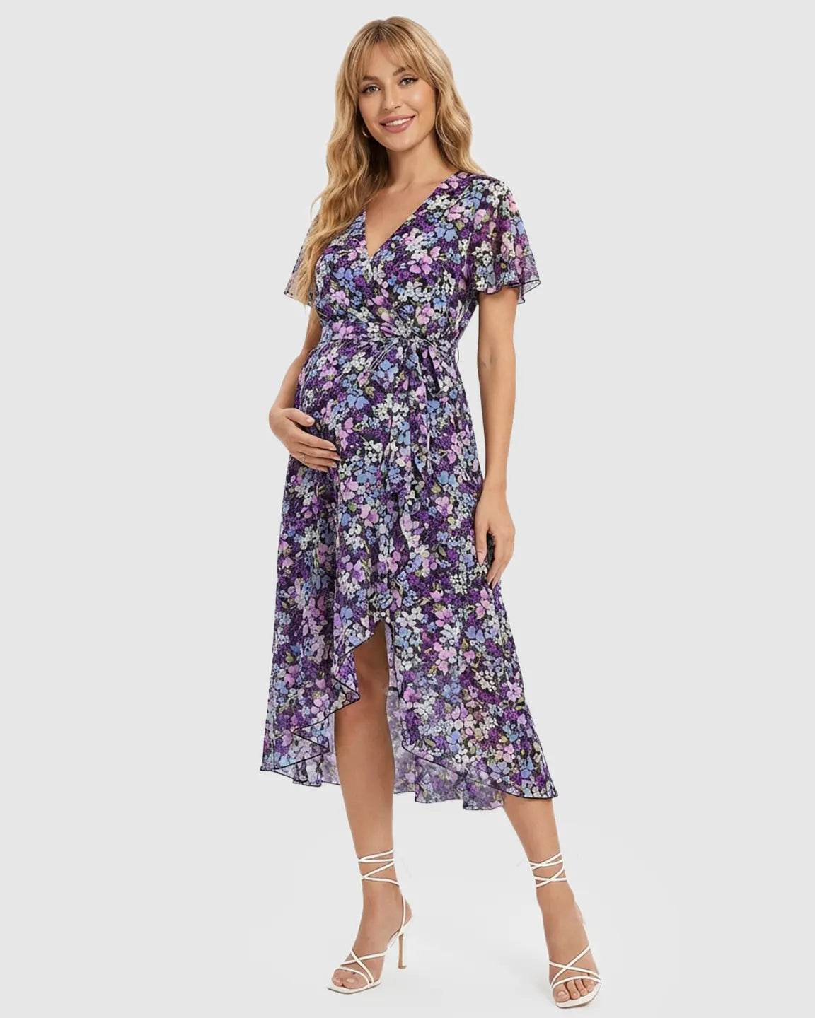Smiling pregnant woman in a purple floral V-neck midi dress with ruffled hem and tie waist.