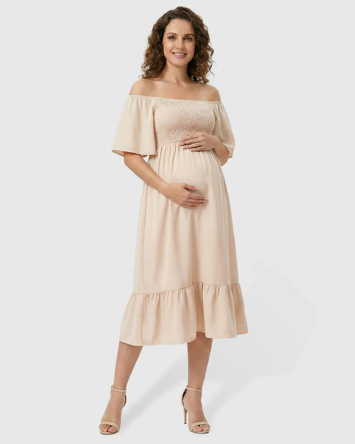 Smiling pregnant woman in a beige off-shoulder smocked maternity midi dress with flutter sleeves and tiered skirt.