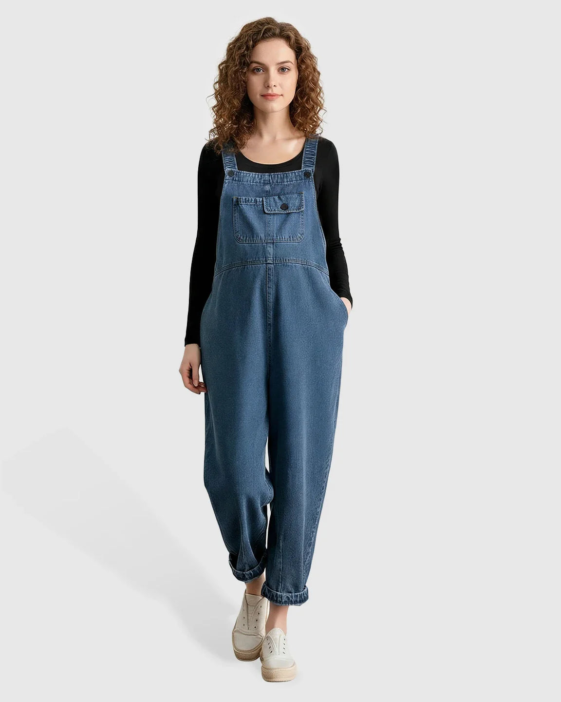 Woman wearing loose-fit blue denim maternity overalls with adjustable straps, chest pocket, and rolled cuffs.