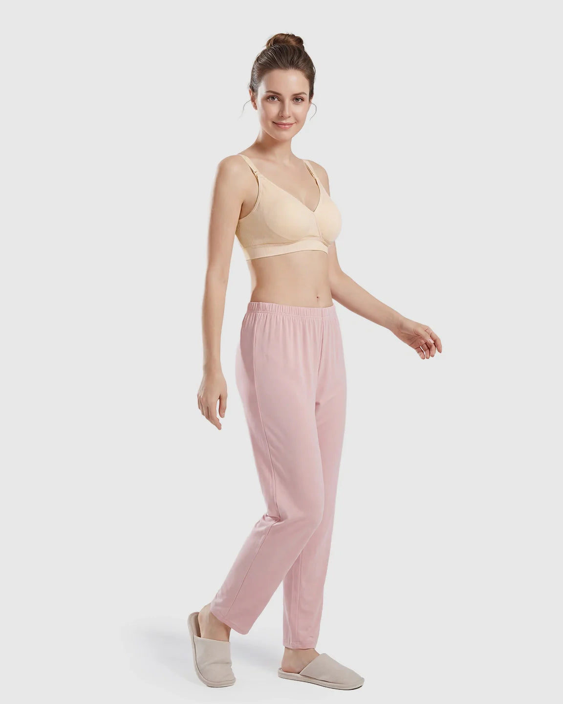 Smiling woman models a seamless, wireless, cross-front beige nursing bra with pink lounge pants and slippers.