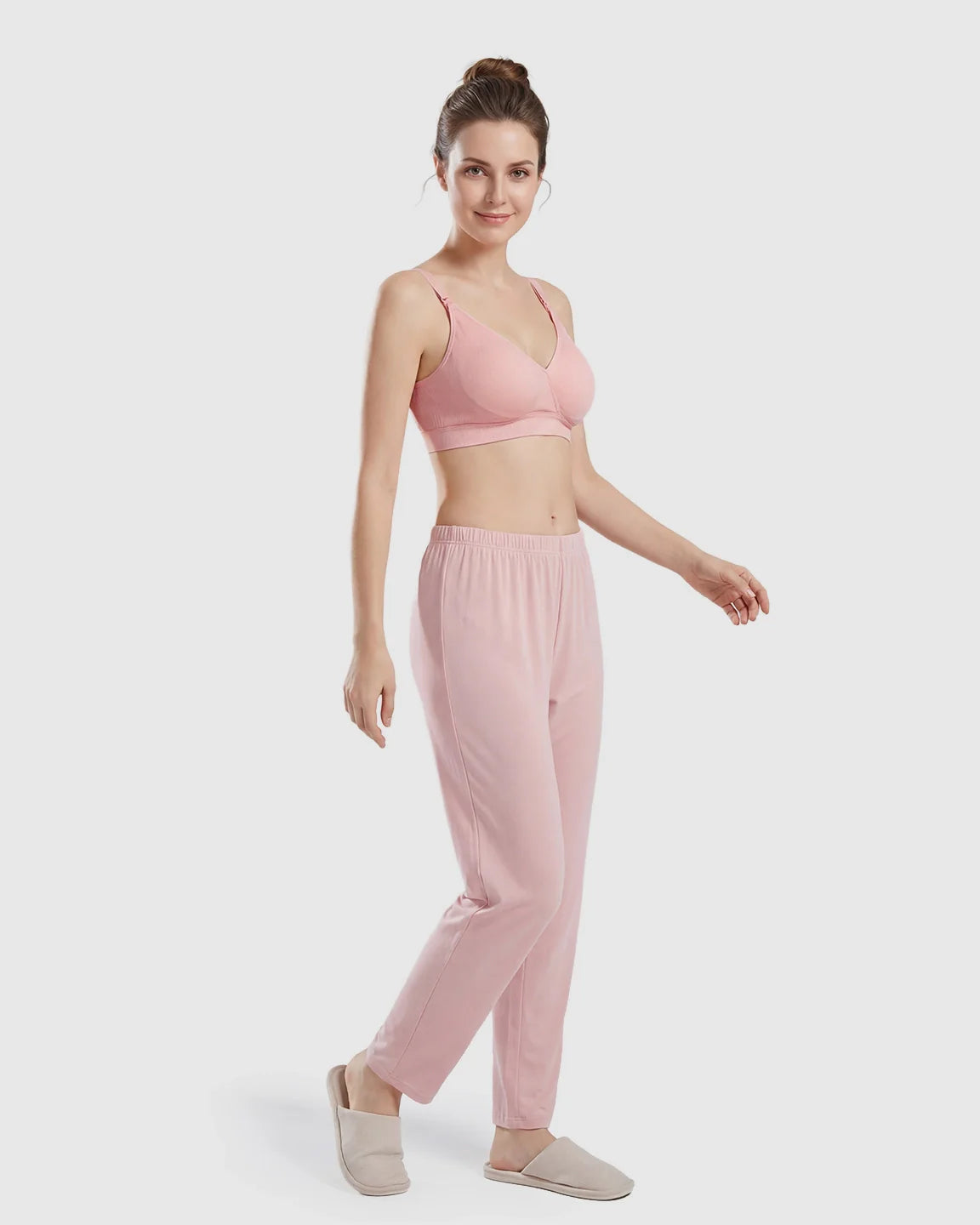 Woman in a pink seamless cross-front wireless nursing bra and matching lounge pants with slippers.