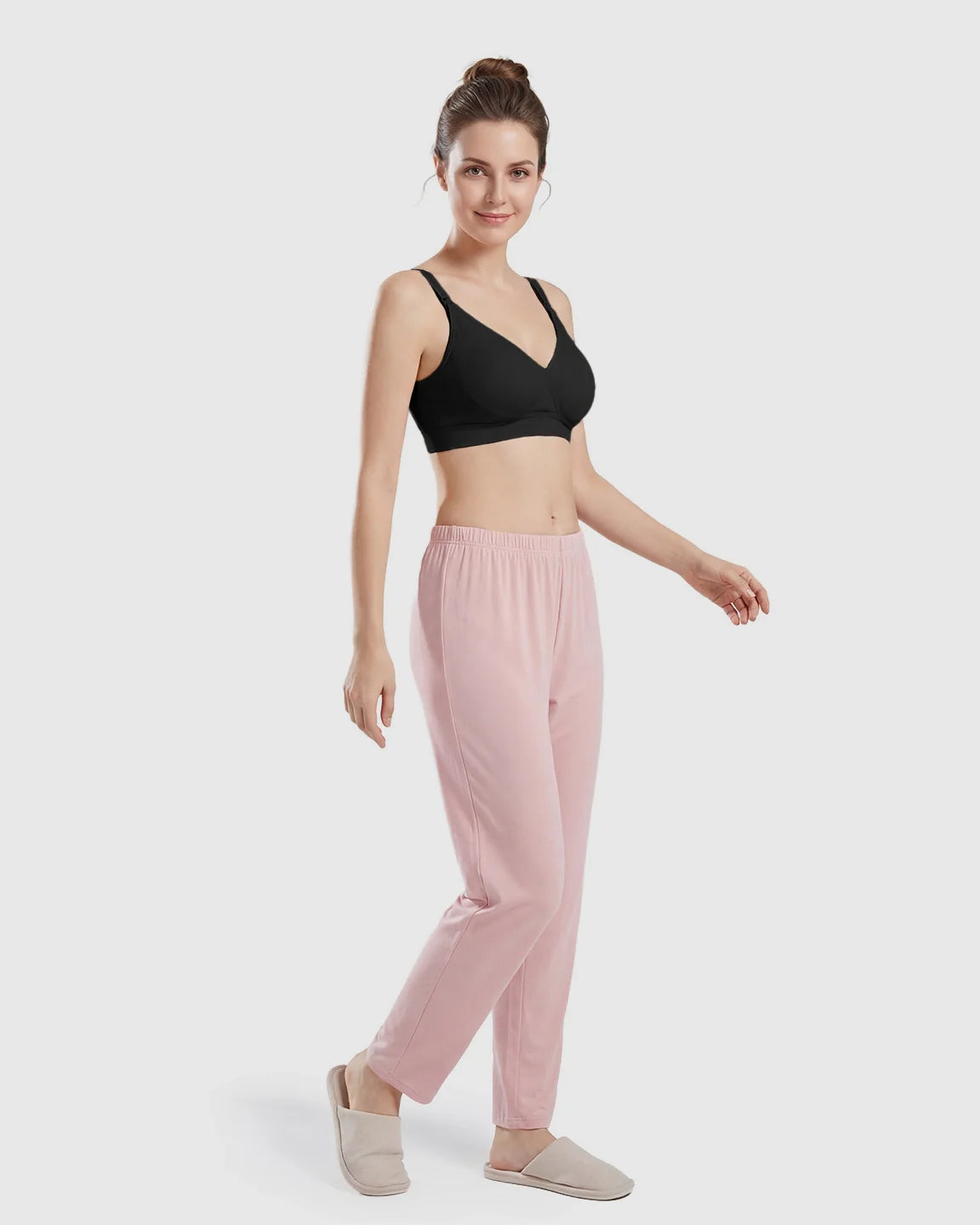 Woman models a black seamless cross-front wireless nursing bra with pink lounge pants and slippers.