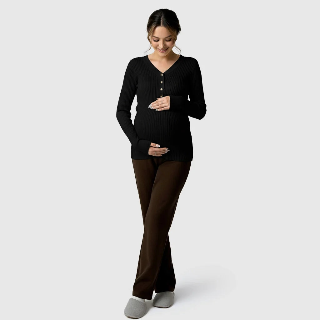 Pregnant woman in a black ribbed V-neck maternity nursing sweater with gold buttons and brown pants.