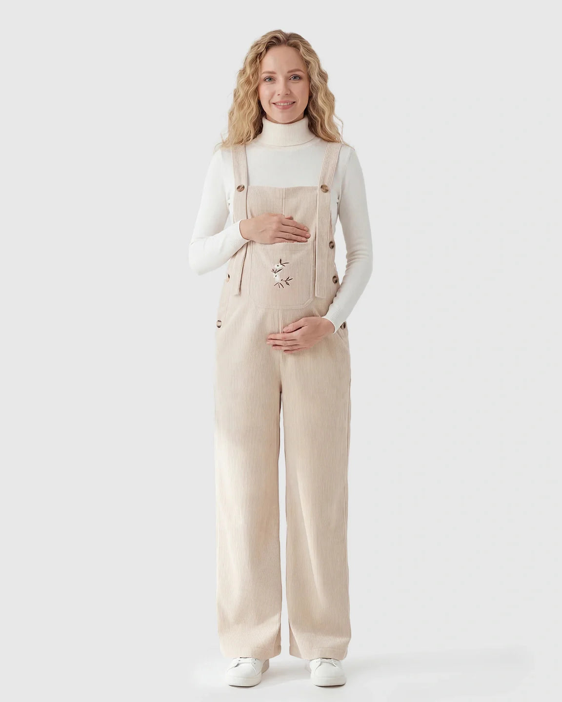 Pregnant woman in beige corduroy maternity overalls with adjustable straps and embroidered pocket.