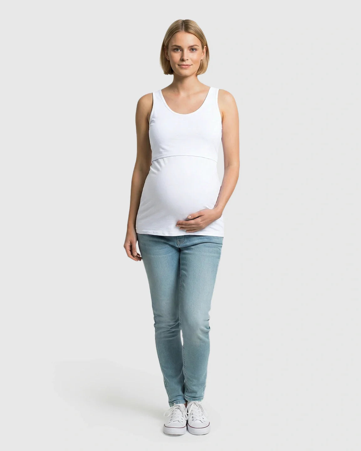 Blonde woman in white stretch nursing-access maternity tank top and jeans, holding her baby bump.