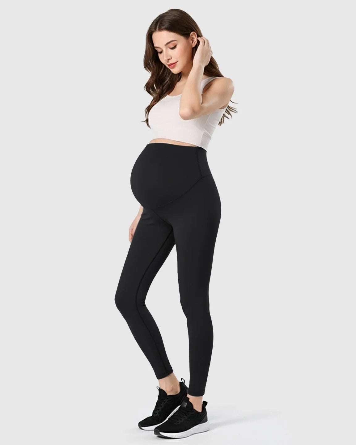 Pregnant woman in black double-layer belly support maternity leggings, white crop top, and black sneakers.