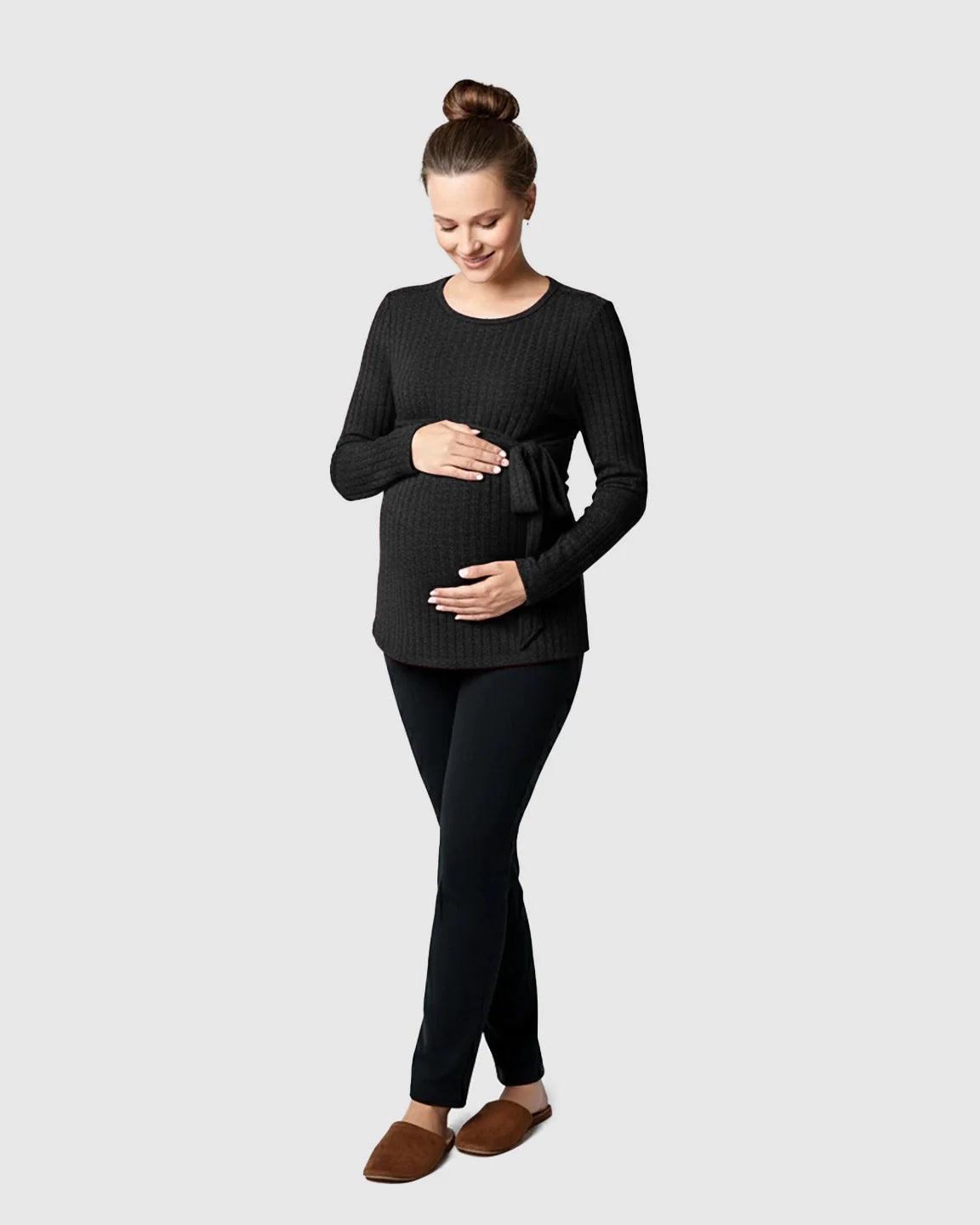Pregnant woman in a black ribbed tie-waist long sleeve maternity sweater, black pants, and brown slippers.