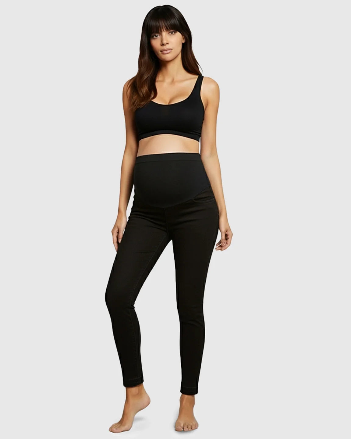 Woman models black high-waist skinny maternity jeans with a supportive elastic waistband and a black bra.