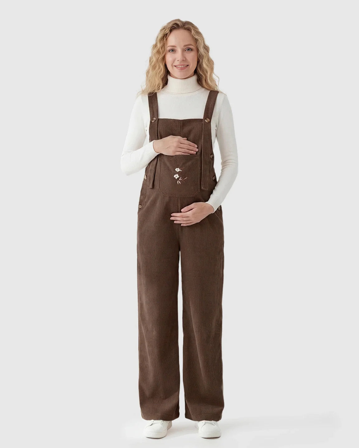 Pregnant woman in brown corduroy maternity overalls with adjustable straps and bird embroidery.