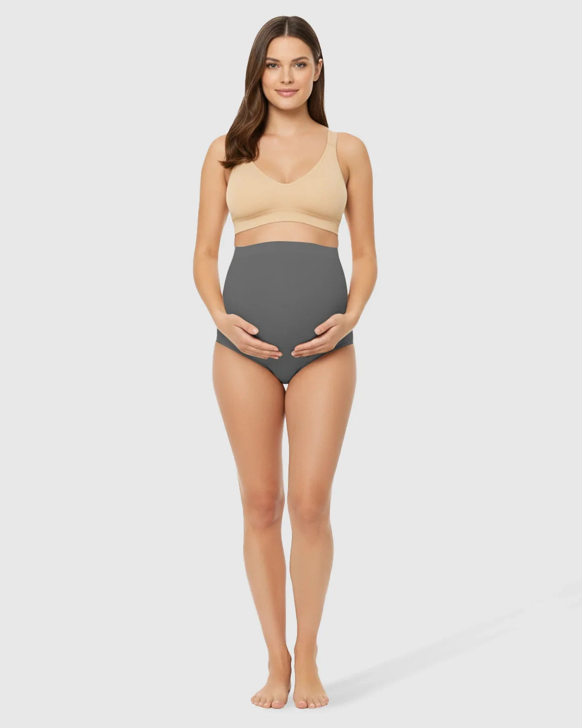Pregnant woman modeling gray high-waist seamless belly-support maternity underwear with a beige bra.