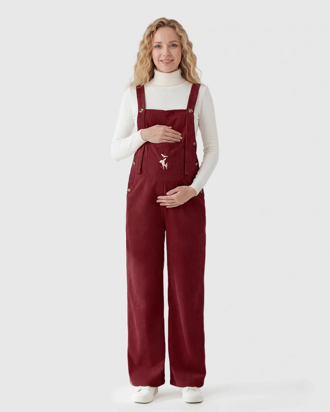 Pregnant woman in red corduroy maternity overalls with adjustable straps and a deer embroidery.