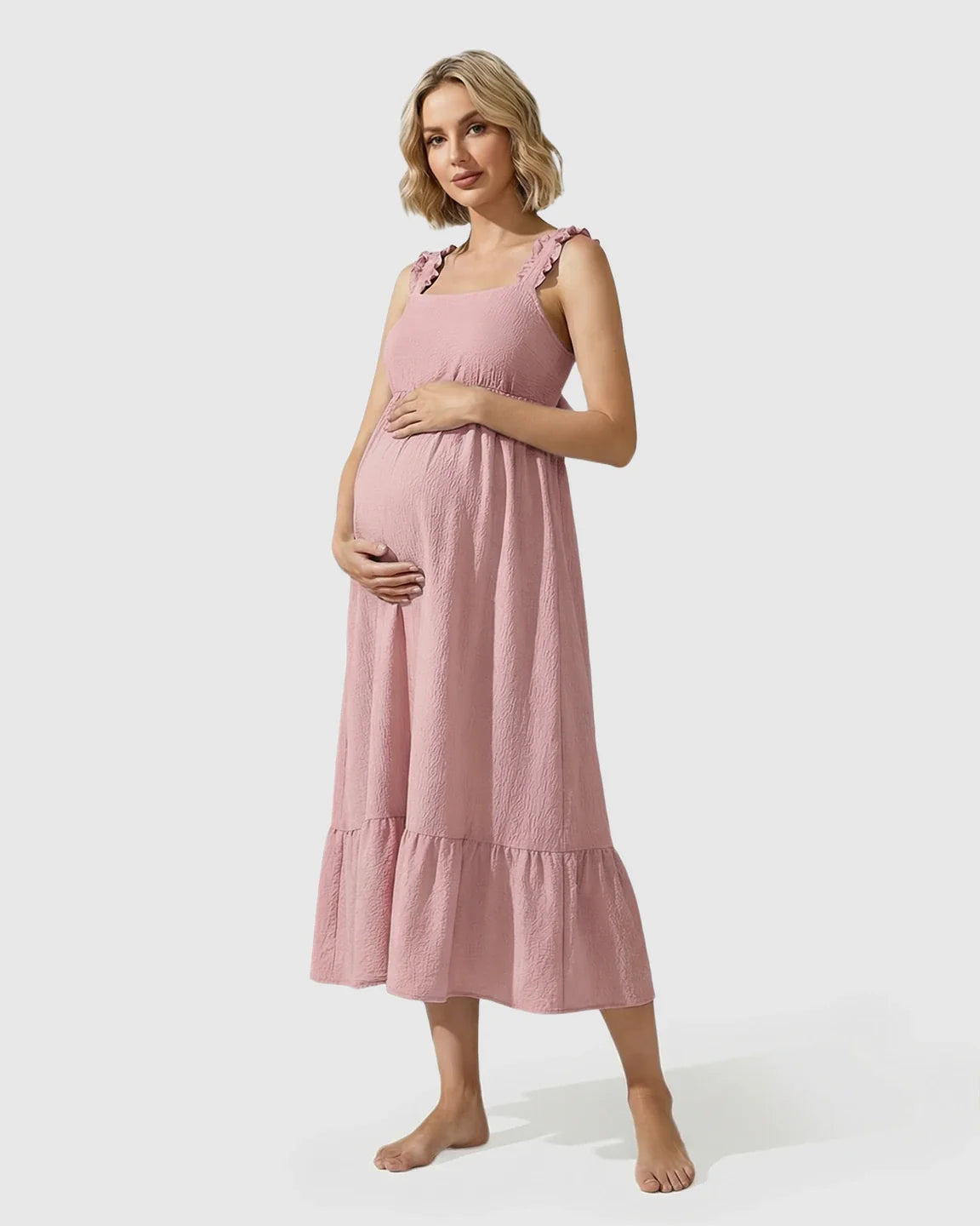 Pregnant woman in a pink ruffled strap tiered maxi maternity dress, holding her baby bump.