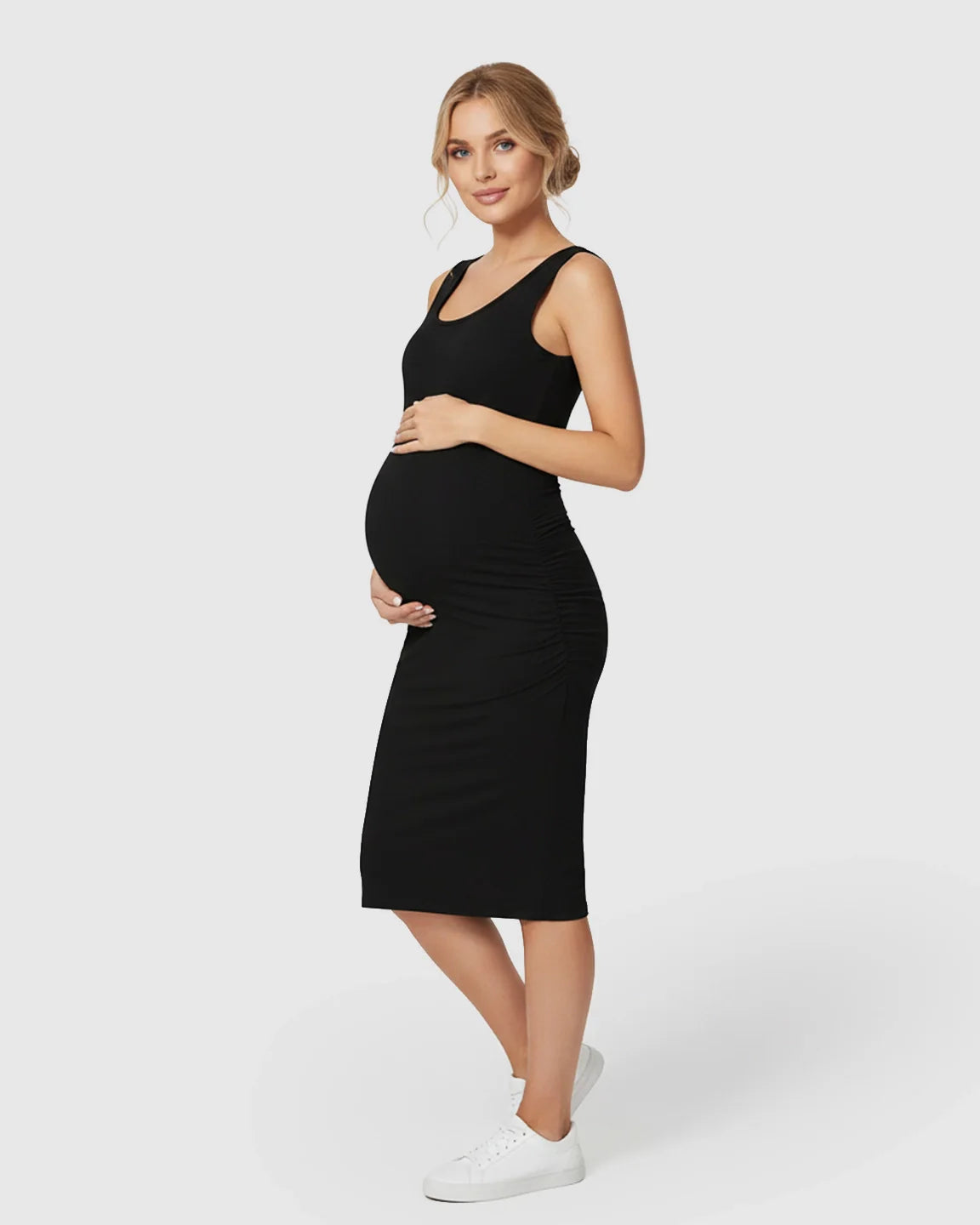 Smiling pregnant woman in a black sleeveless bodycon maternity dress with side ruching, holding her belly.