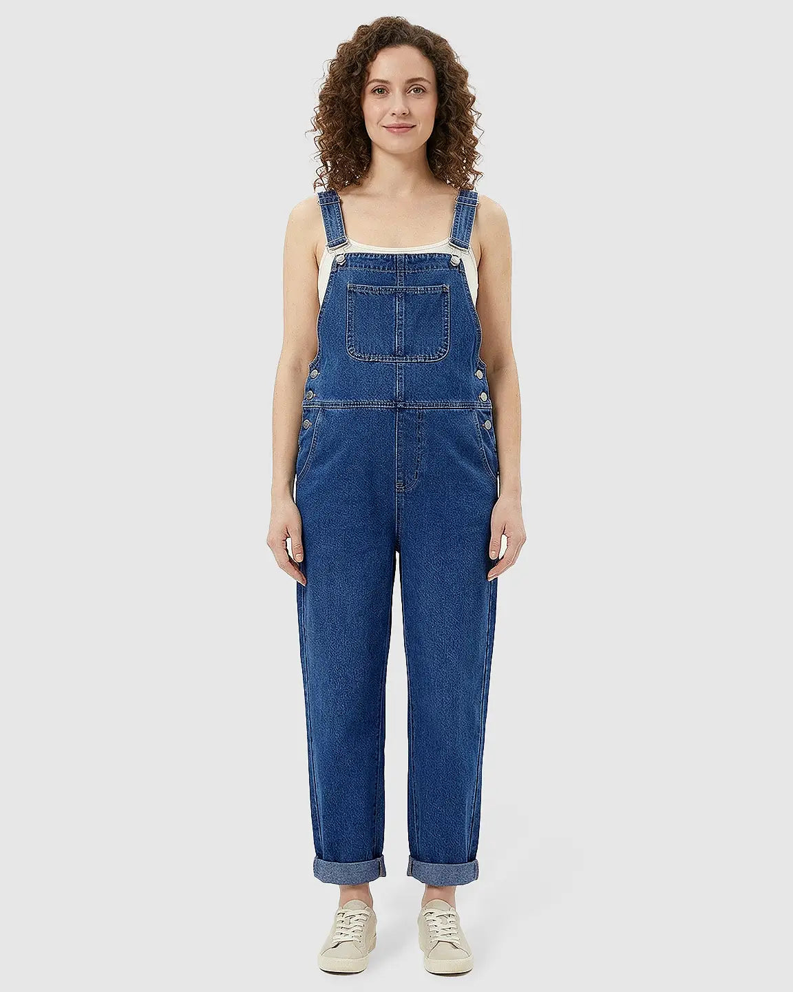 Woman in a blue denim maternity jumpsuit with adjustable straps, front pocket, and cuffed ankles.