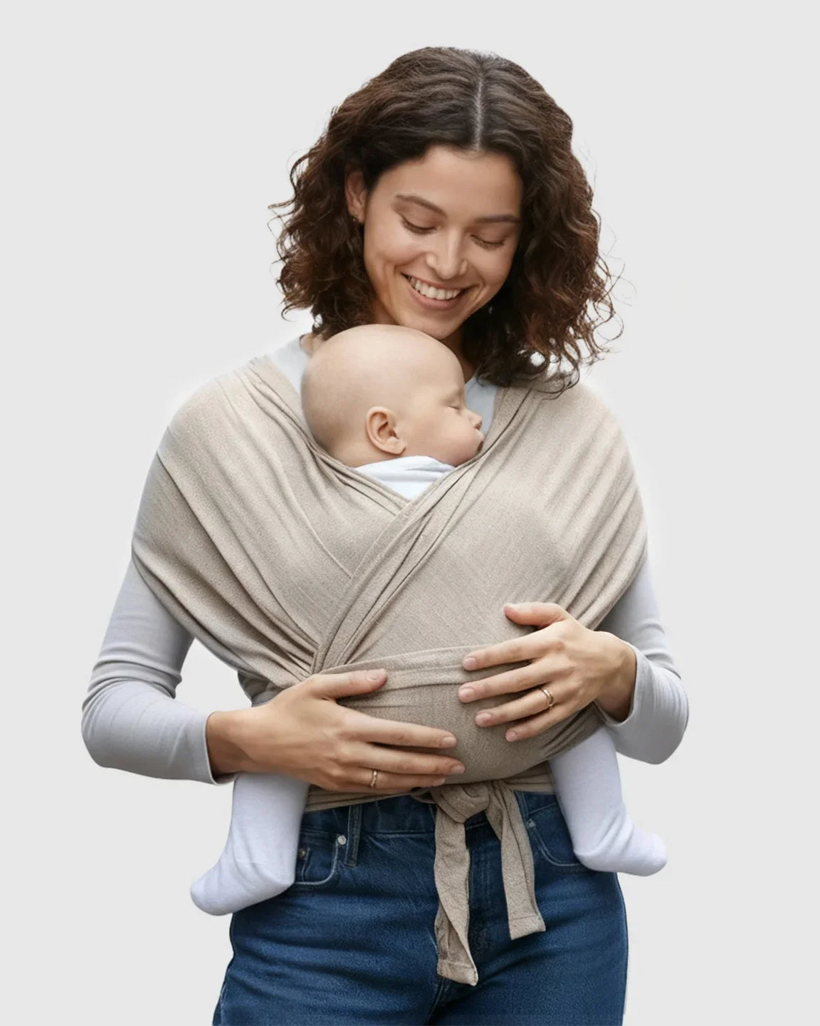 Smiling woman wearing a beige adjustable wrap-around baby carrier with a sleeping infant.