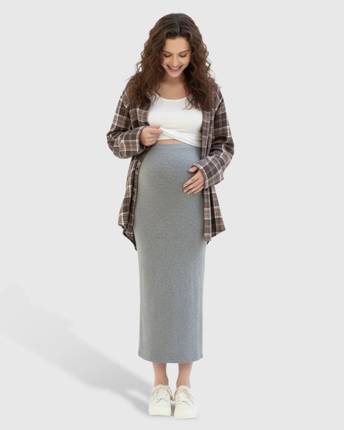 Smiling pregnant woman in a gray midi skirt, white top, and brown plaid shirt, gently touching her bump.