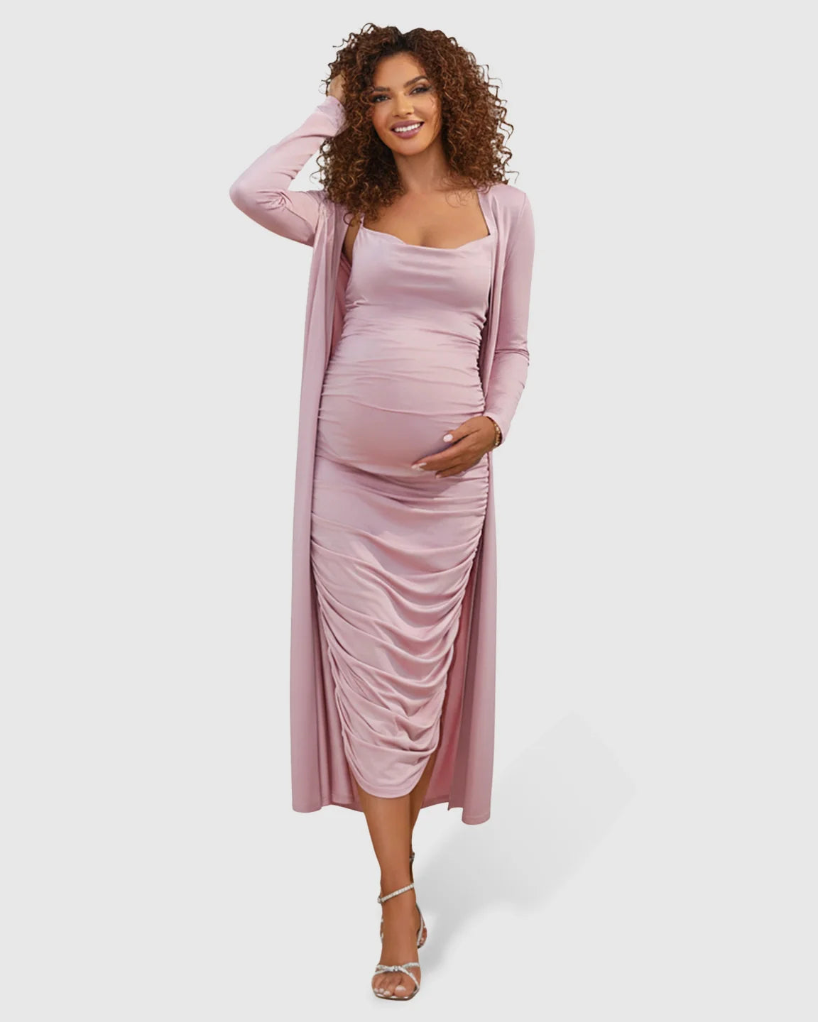 Smiling pregnant woman in a pink side-ruched slip dress and long-sleeve cardigan maternity set.