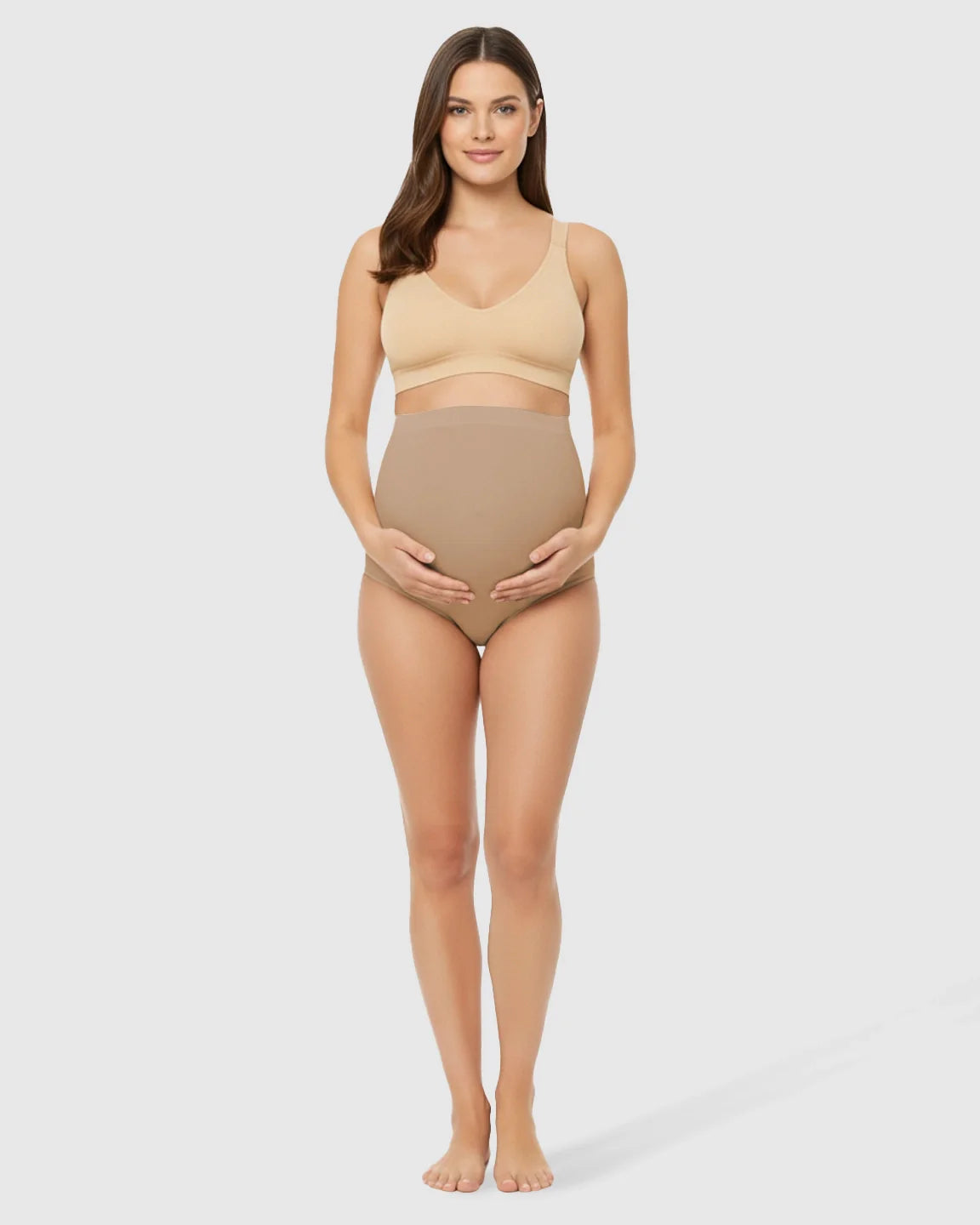 Woman wearing seamless nude high-waist belly-support maternity underwear and matching bra, cradling her bump.