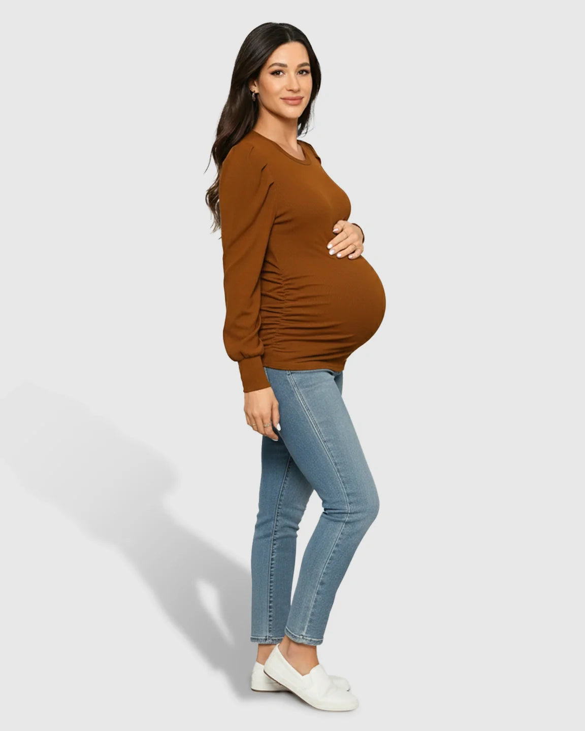 Maternity model in a warm brown long-sleeve ruched top and jeans, cradling her bump.