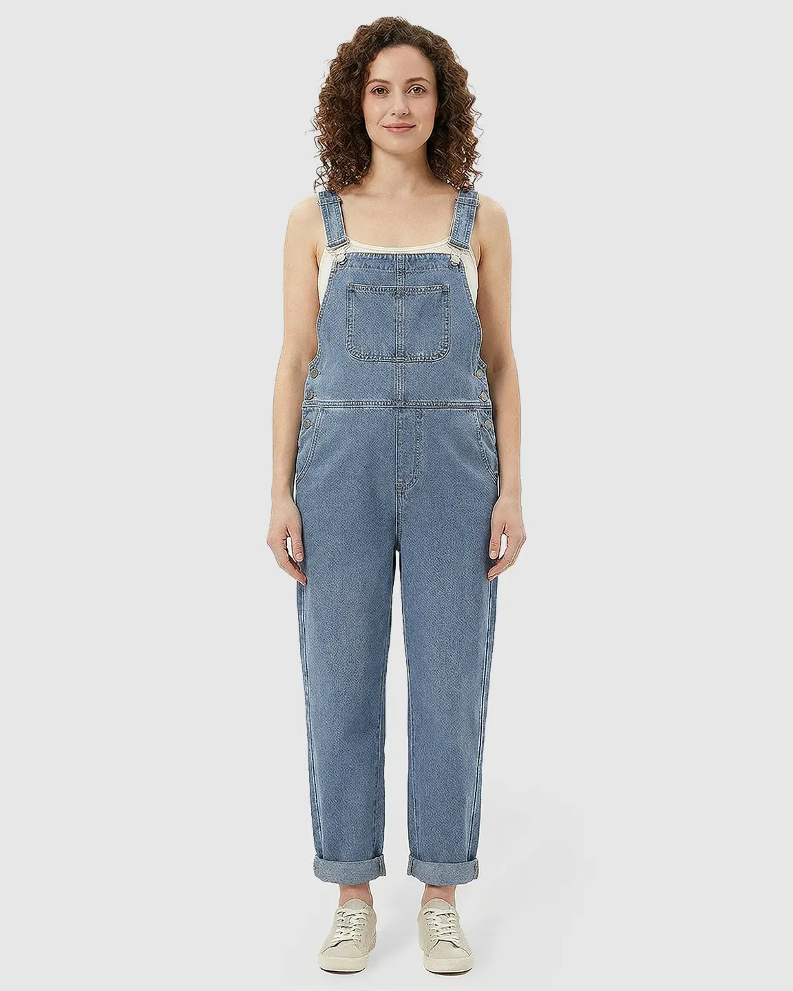 Woman in a light wash blue denim maternity jumpsuit with adjustable straps, chest pocket, and rolled cuffs.