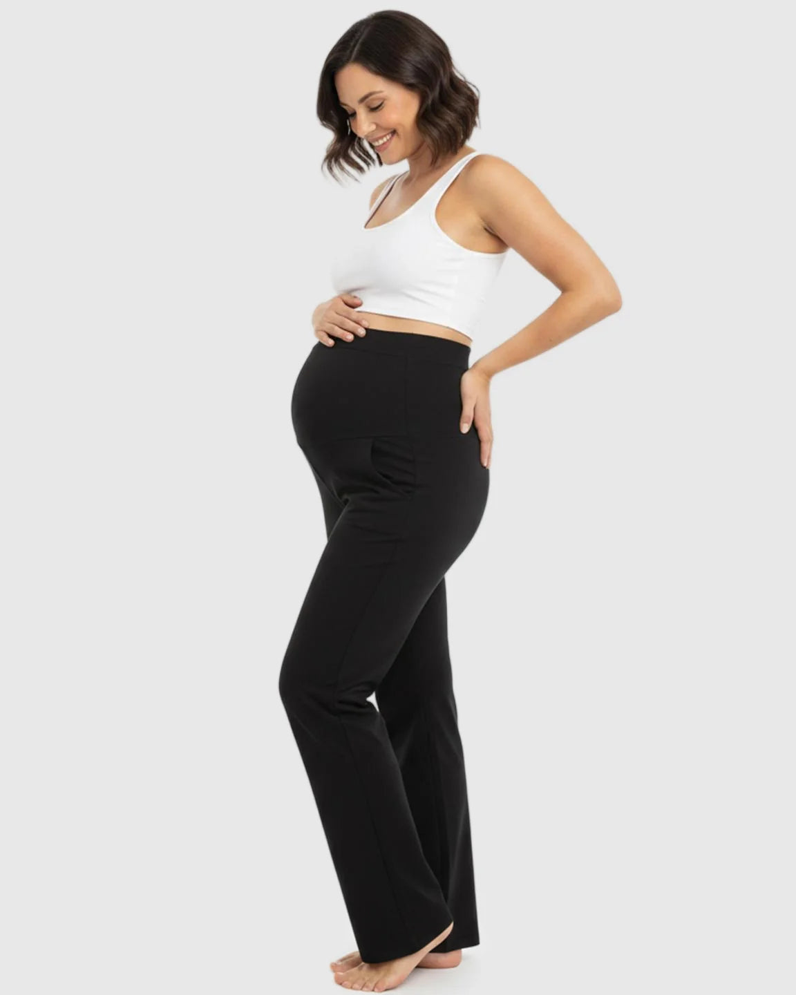 Smiling pregnant woman in black stretch flared maternity pants with an over-belly waistband and a white crop top.