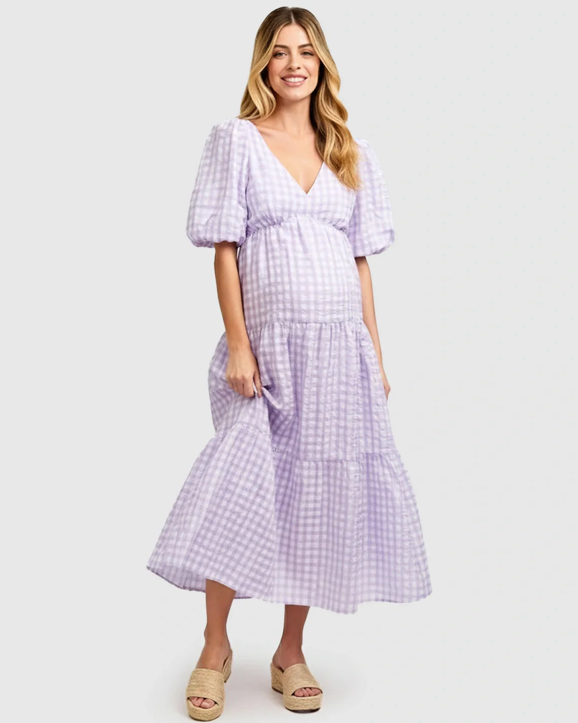 Smiling pregnant woman in a lavender gingham puffed sleeve tiered maxi dress and espadrille sandals.