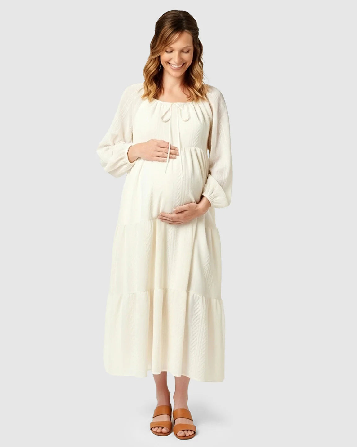 Pregnant woman in a cream textured long-sleeve tiered maternity midi dress with a tie-front neckline.