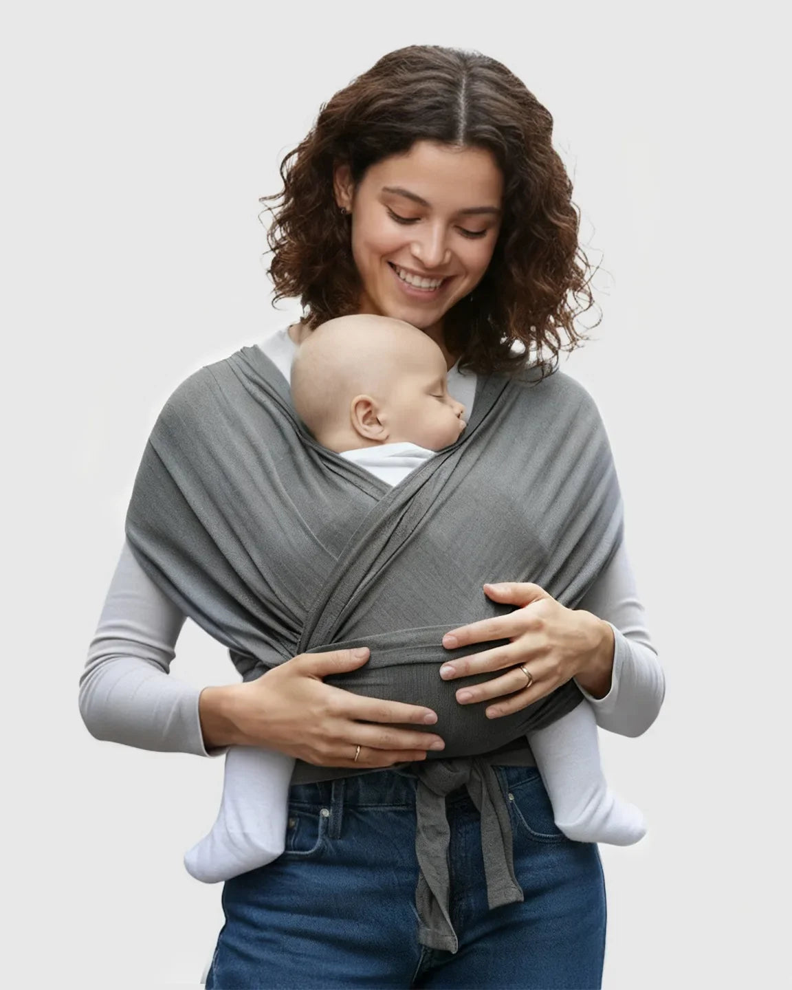 Smiling woman wearing a gray, soft, adjustable wrap-around baby carrier with a sleeping infant.