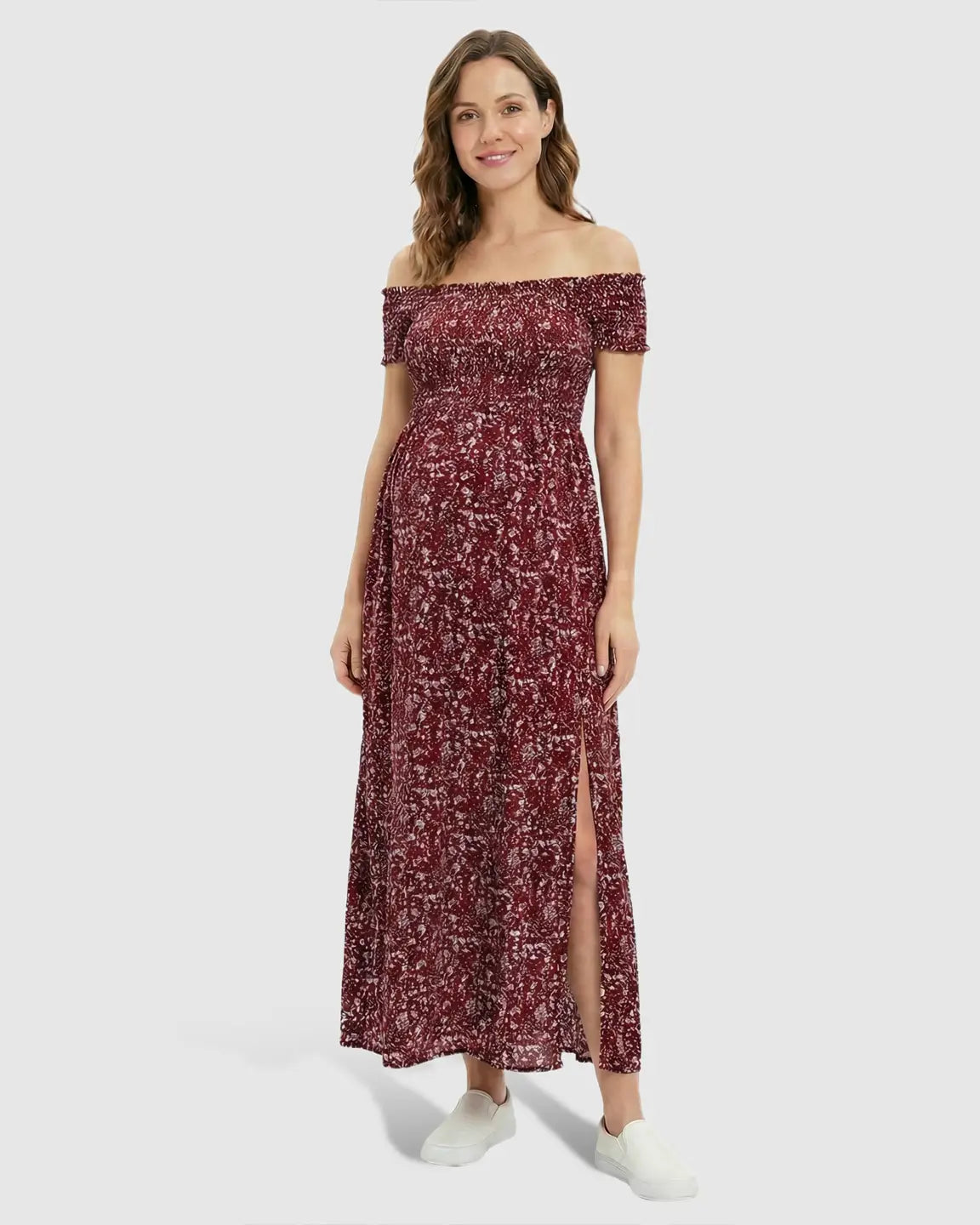 Smiling pregnant woman in a red floral off-shoulder maxi dress with a side slit and white sneakers.