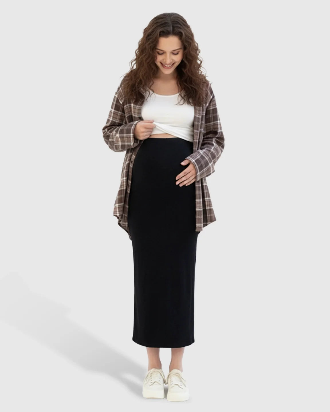 Smiling pregnant woman in a black maternity midi skirt, white top, and brown plaid shirt.