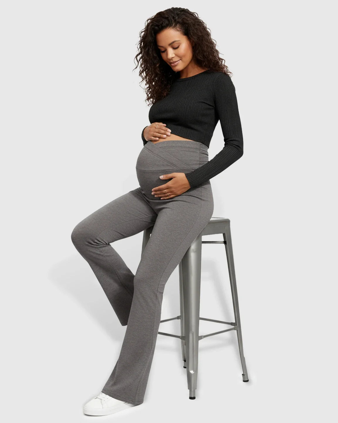 Pregnant woman in grey Bump Society crossed waistband flared maternity pants, black top, and white sneakers.