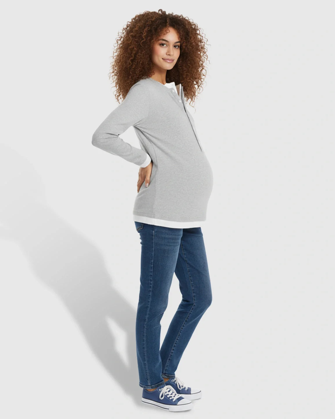 Pregnant woman in a gray long-sleeve henley maternity and nursing top with white trim, blue jeans, and sneakers.