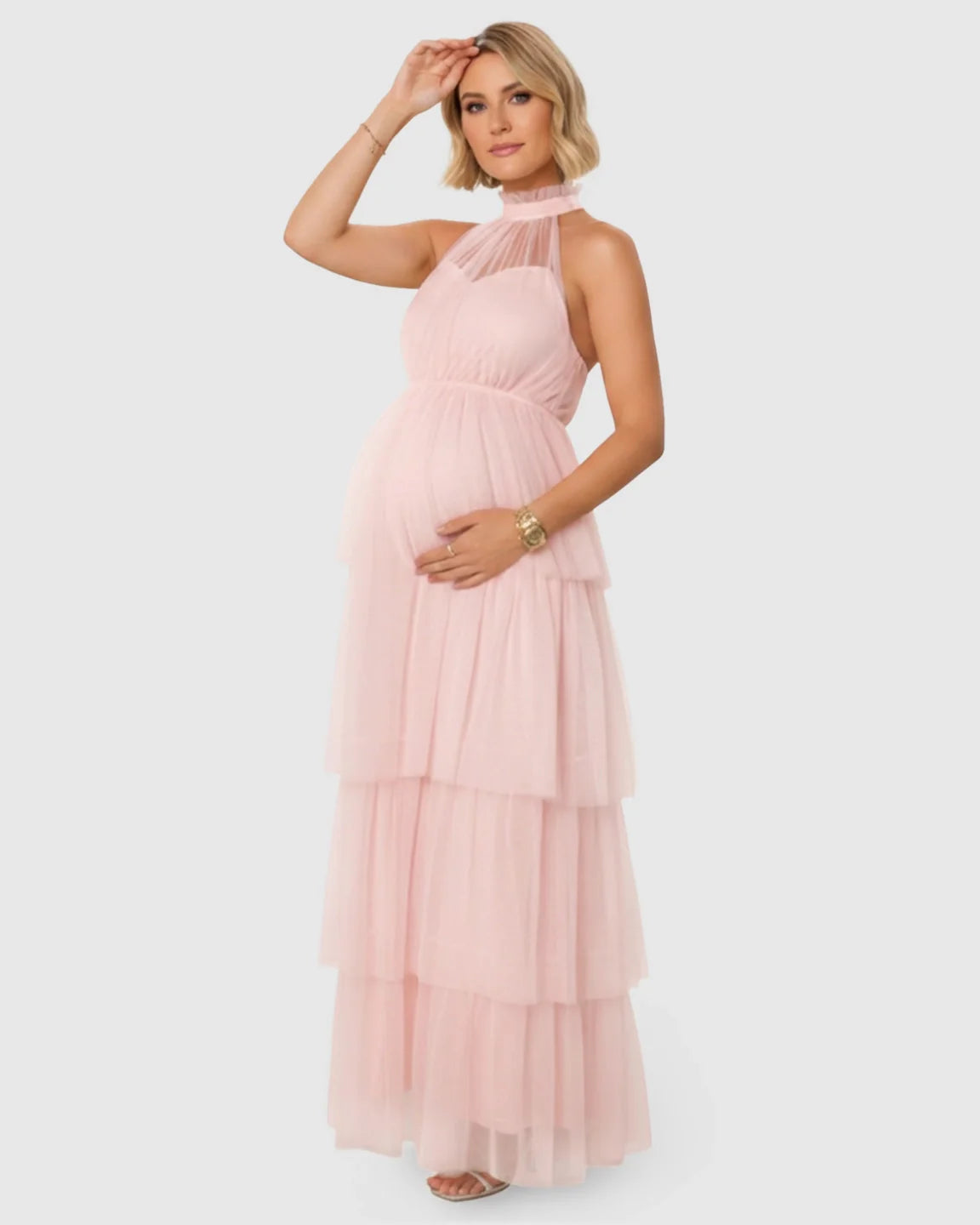 Blonde pregnant woman in a pink halter neck tiered mesh maternity gown, gently touching her bump.