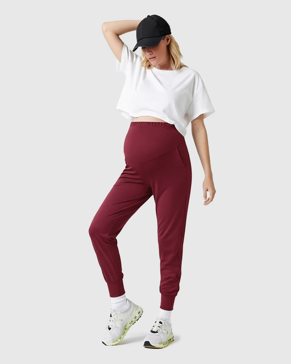 Woman in supportive slim-fitting maroon maternity joggers with high-waist belly support, white crop top, and black cap.