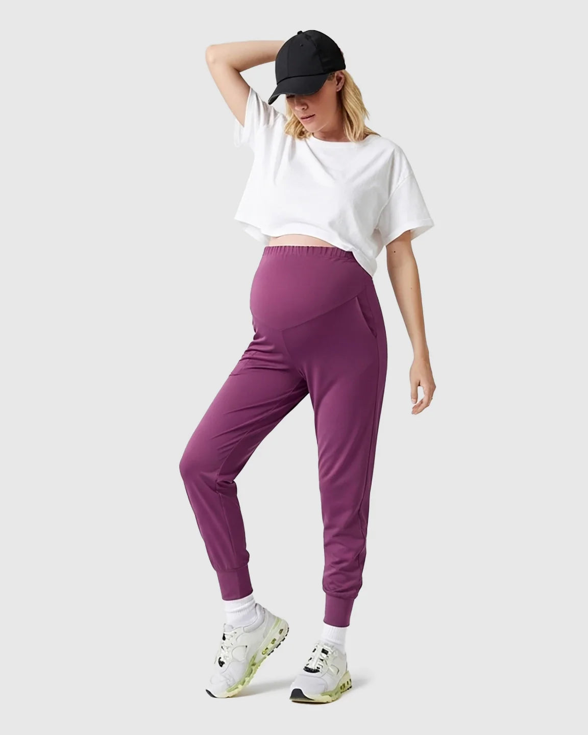 Woman in purple supportive slim-fitting maternity joggers with a high-waist design, white crop top, and black cap.