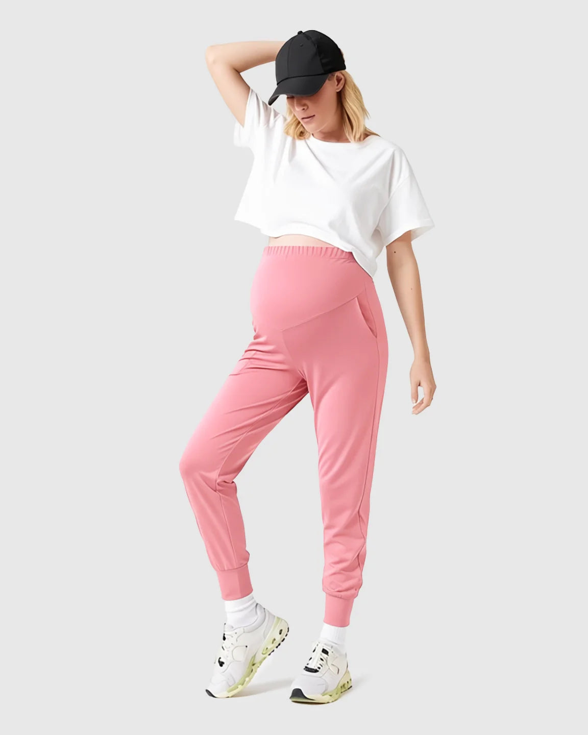 Woman in pink supportive maternity joggers, white crop top, black cap, and sneakers.