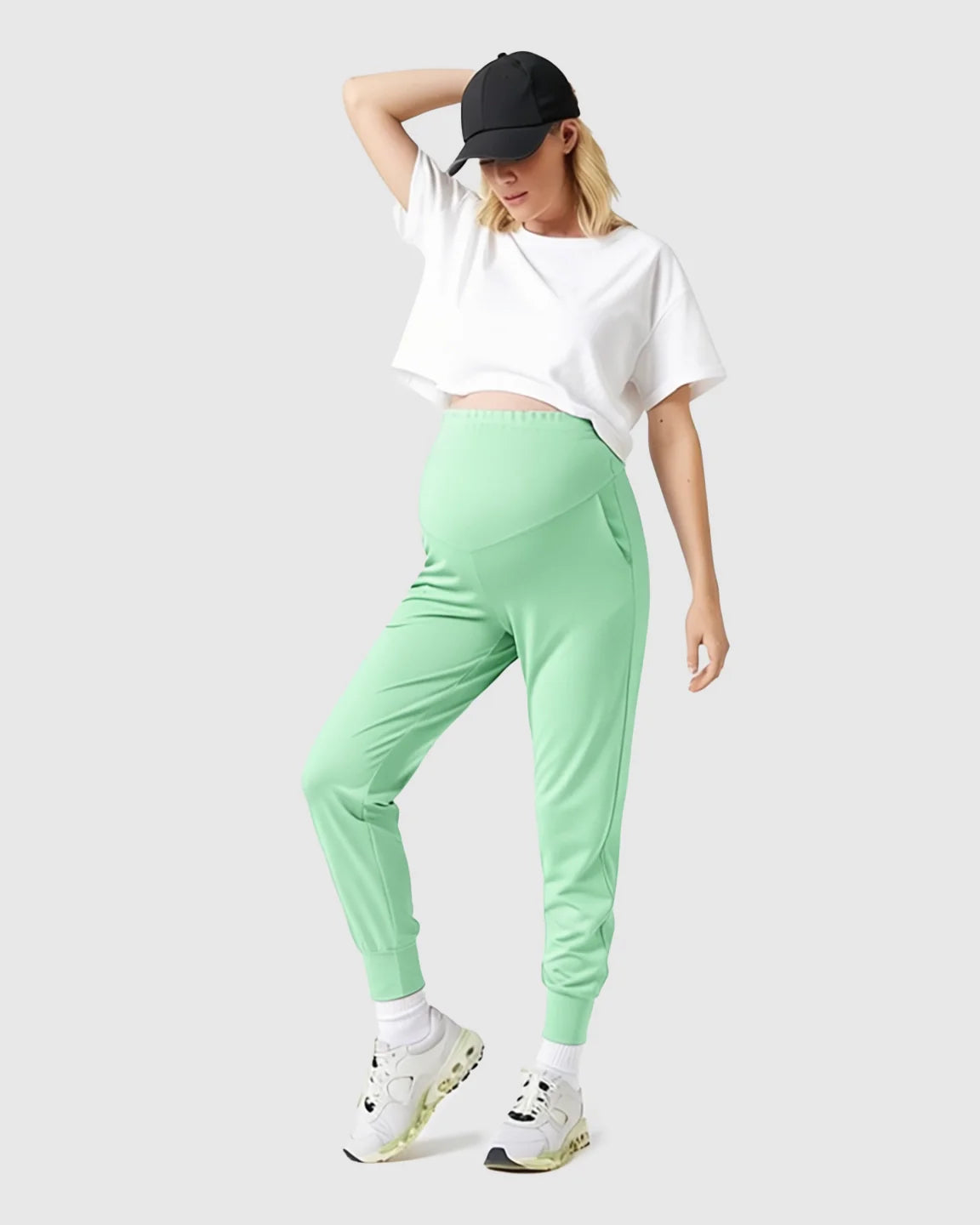 Woman in mint green supportive maternity joggers with a high-waist design, white crop top, and black cap.