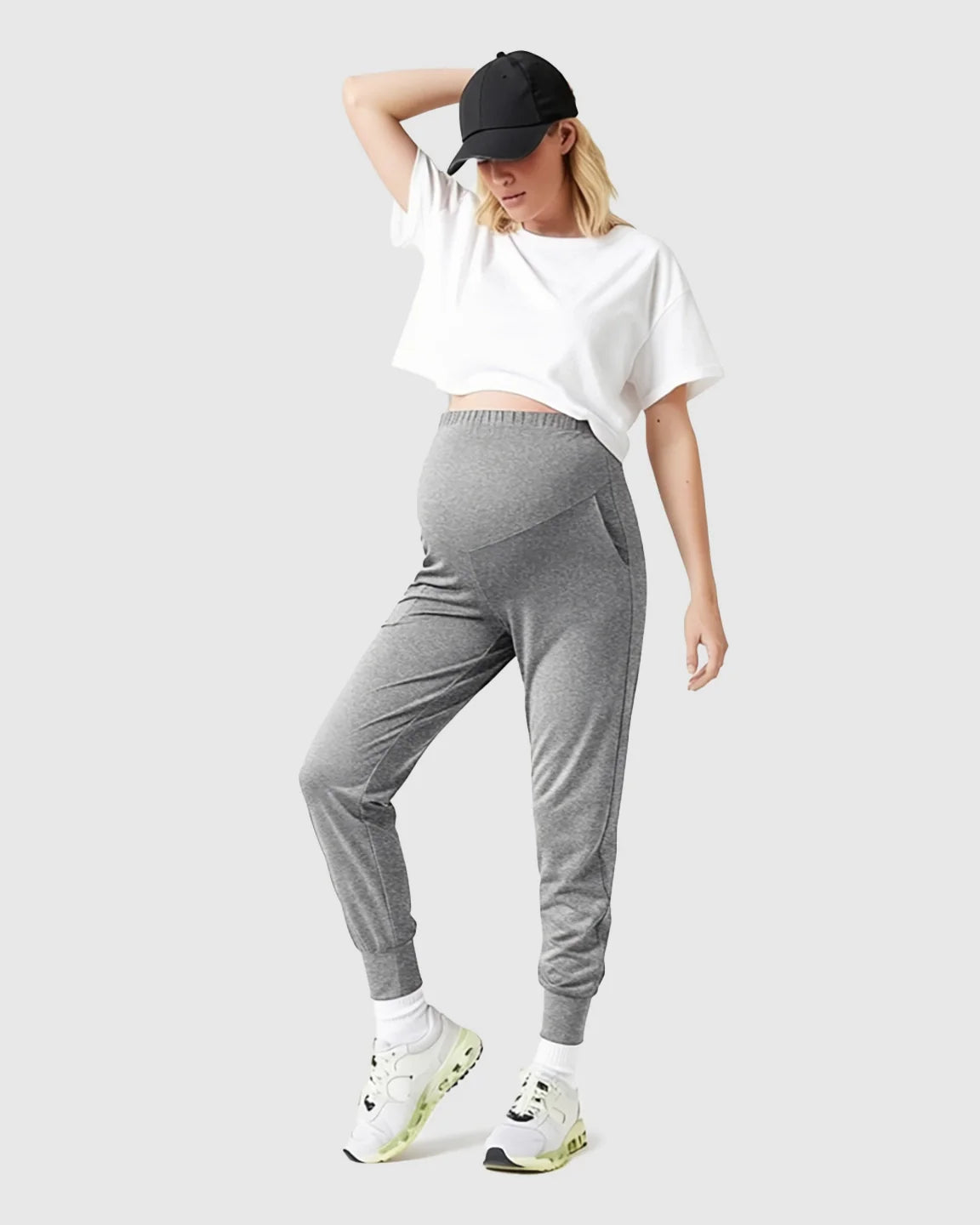 Woman in grey supportive slim-fitting maternity joggers with a high-waist belly panel, white crop top, and black cap.